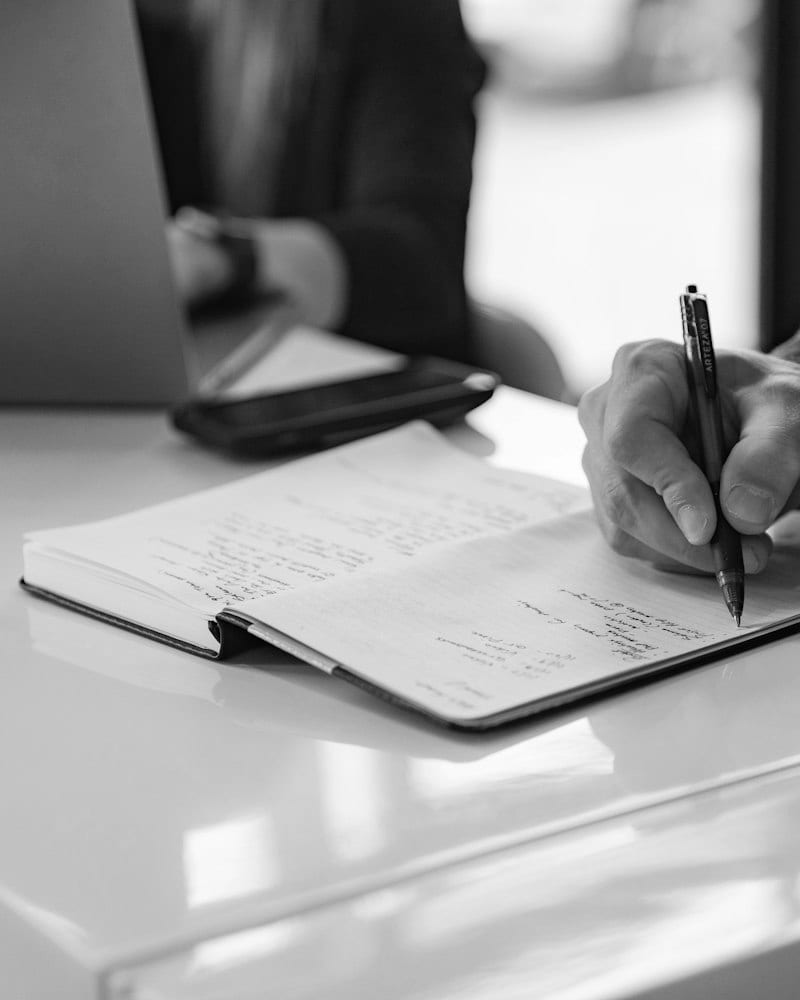 A hand writing in a notebook at a bright desk with a blurred figure in the background