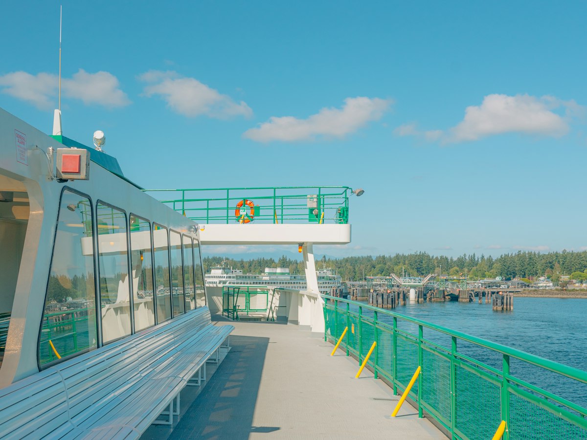 Empty ferry deck with green railings, wooden benches, and a small pier in the distance